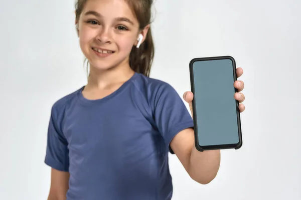 Pequeña niña deportiva con auriculares, sonriendo a la cámara y mostrando el teléfono inteligente con la pantalla en blanco, de pie aislado sobre el fondo blanco — Foto de Stock