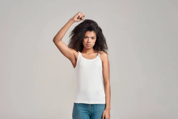 Retrato de jovem afro-americana com cabelo encaracolado vestindo camisa branca com sério olhar confiante ao mostrar, levantando o punho cerrado, de pé isolado sobre fundo cinza — Fotografia de Stock