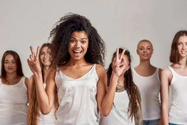 Portrét mladé Afroameričanky v bílém tričku s mírovým znamením před kamerou. Skupina různých žen stojící izolované nad šedým pozadím — Stock fotografie