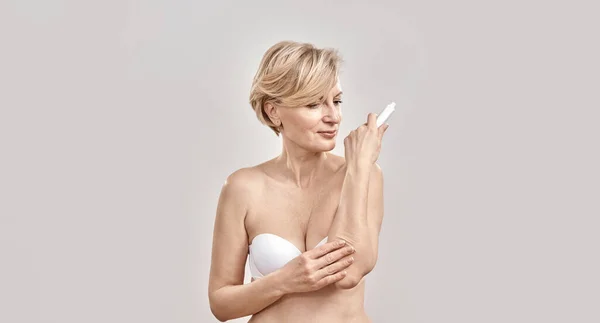 Attractive middle aged woman applying exfoliating moisturizing cream on dry elbow skin while posing isolated over grey background — Stock Photo, Image