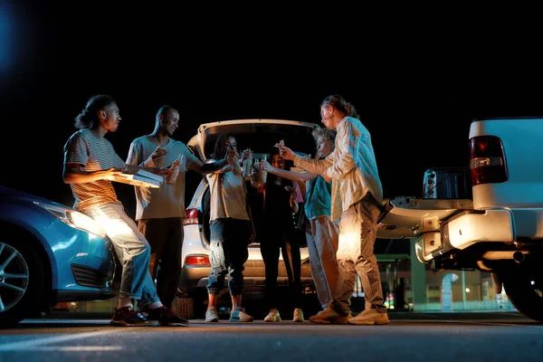 Jóvenes amigos casualmente vestidos de diferentes nacionalidades de pie uno frente al otro afuera en un sitio de estacionamiento sacudiendo botellas de cerveza mientras comen pizza — Foto de Stock