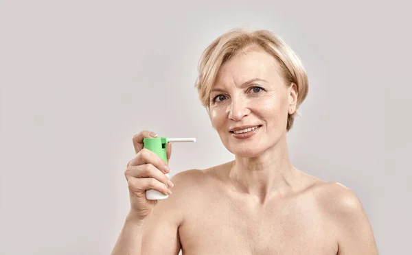 Portrait of attractive middle aged woman smiling at camera and holding throat spray, standing isolated over grey background — Stock Photo, Image