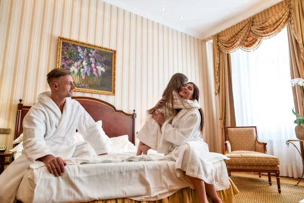 Disfruta de la estancia. Los padres caucásicos y su hija en batas blancas relajarse y pasar la mañana juntos. Están sentados en la cama en una lujosa habitación de hotel. Familia, viajes, resort, concepto de vacaciones — Foto de Stock