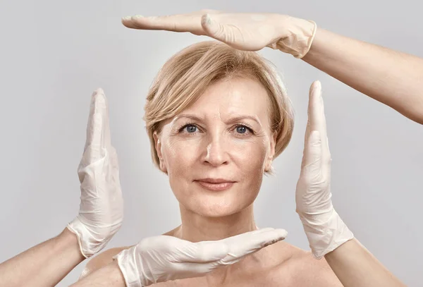 Portrait of attractive middle aged woman looking at camera. Beautician creating a frame around female face with hands in gloves isolated against grey background — Stock Photo, Image