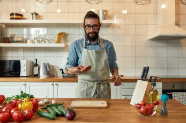 Genç adam, İtalyan aşçı önlüklü kameraya bakıyor, bıçağın ucunu tutuyor ve mutfakta sebzelerle sağlıklı bir yemek hazırlıyor. Ev konseptinde aşçılık