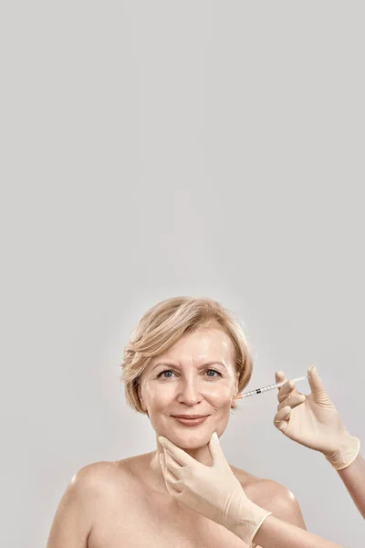 Portrait of beautiful naked middle aged woman looking at camera while she gets injection in her face, posing isolated against grey background — Stock Photo, Image