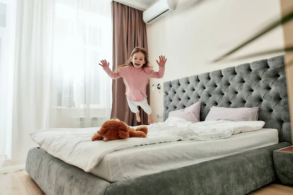 Мила маленька дівчинка розважається і стрибає вниз, як маленький тигр на її коричневий плюшевий ведмідь босоніж на ліжку — стокове фото