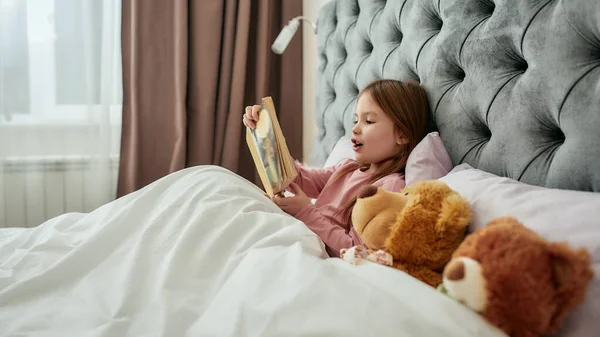 Uma menina fofa segurando seu livro de perto e gostando de ler em voz alta, deitada em uma cama grande com seus teddybears em um quarto espaçoso brilhante em casa — Fotografia de Stock
