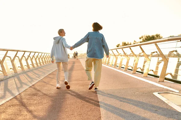 Full length shot of happy lesbian couple holding hands, walking on the bridge and watching the sunrise together. Homosexuality, LGBT and love concept — Stock Photo, Image