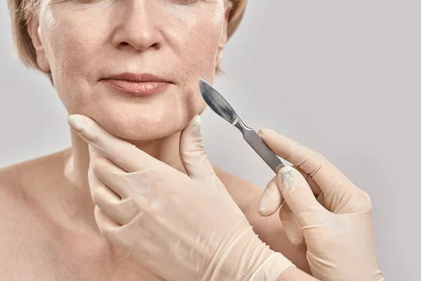 Ujęcie twarzy kobiety w średnim wieku i skalpel medyczny w rękach lekarzy odizolowanych na szarym tle — Zdjęcie stockowe