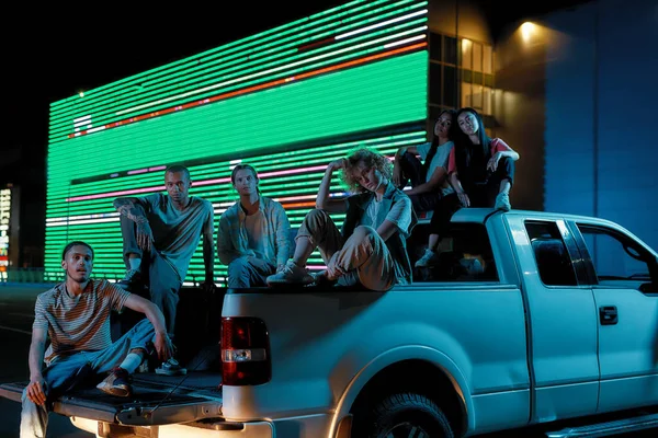 A group of six good-looking casually dressed youngsters of different nationalities sitting in an opened pick-ups trunk next to each other posing on a camera with a led screen behind — Stock Photo, Image