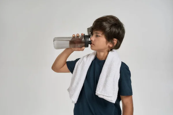 Mały sportowy chłopiec w odzieży sportowej pijący wodę z butelki stojąc z białym ręcznikiem na szyi, odizolowany na białym tle. — Zdjęcie stockowe