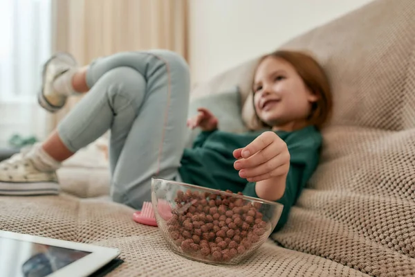 Uma menina bonita sentada perna a perna em um sofá assistindo TV comendo bolas de cereais de uma tigela vítrea em uma grande sala brilhante — Fotografia de Stock