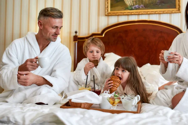 Melhor pequeno-almoço. Pais e duas crianças em roupões de banho brancos tomando café da manhã na cama, comendo doces e bebendo café no luxuoso quarto de hotel. Família, resort, conceito de serviço de quarto — Fotografia de Stock