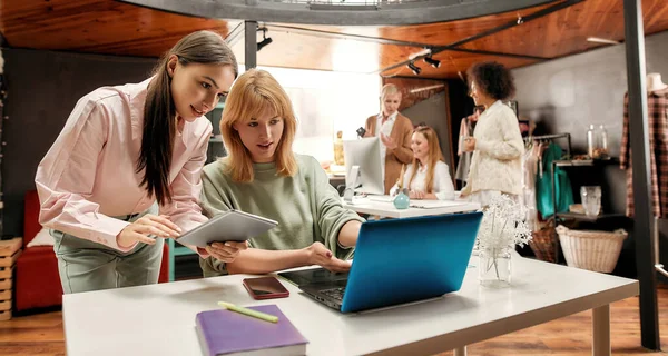 Zwei gut gekleidete weiße junge Frauen, die vor einem Laptop miteinander reden, und ihre Mitarbeiterinnen im Hintergrund — Stockfoto