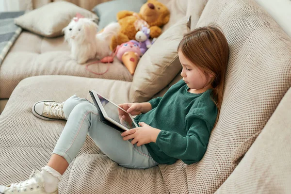 En liten flicka som sitter på en soffa och leker lekar på en surfplatta med skorna på — Stockfoto