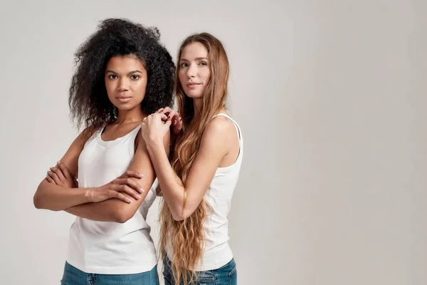 Ritratto di due giovani donne diverse che indossano camicie bianche guardando la macchina fotografica mentre posano insieme isolate su sfondo grigio — Foto Stock