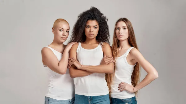 Drie zelfverzekerde jonge diverse vrouwen met witte shirts die naar de camera kijken terwijl ze samen poseren geïsoleerd over een grijze achtergrond — Stockfoto