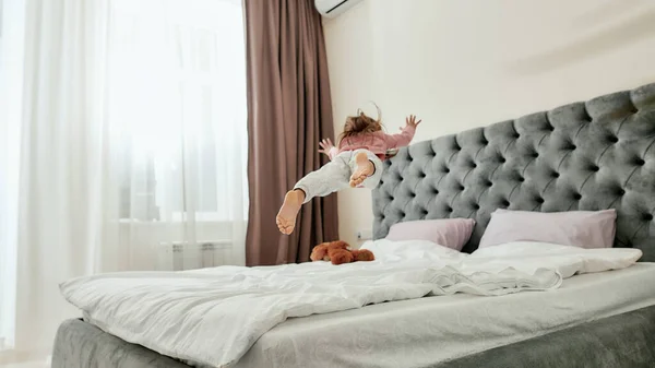 Seorang gadis kecil melompat-lompat dan jatuh seperti bintang di tempat tidur bertelanjang kaki dengan boneka beruang — Stok Foto