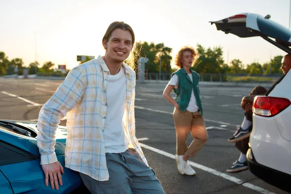 Een gewone gekleed langharige blanke man kijkt in een camera glimlachend tegen een auto buiten op een parkeerplaats met zijn vrienden op een achtergrond — Stockfoto