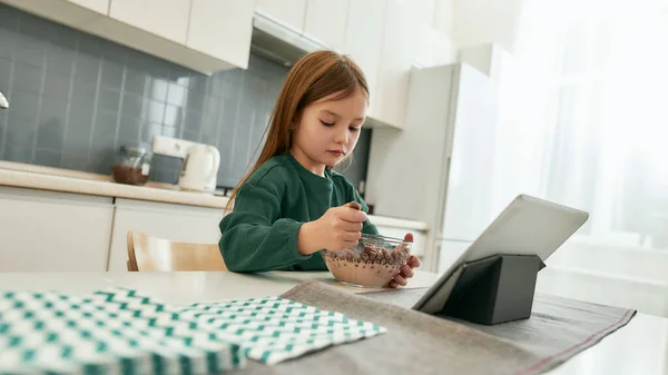 Uma menina pequena está sentada à mesa em uma grande cozinha brilhante olhando para uma tigela cheia de leite e bolas de cereais — Fotografia de Stock