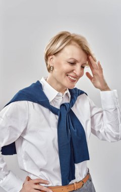 Kafkasyalı zarif bir orta yaşlı kadının portresi iş elbisesi giymiş, gülümseyerek kafasına dokunuyor, gri arka planda soyutlanmış bir şekilde poz veriyor.