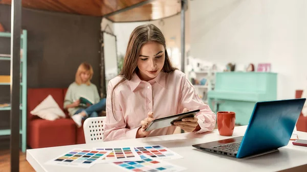 若いですかわいいです茶色のストレート髪の女性よく服を着た彼女のタブレットと同僚とテーブルの上に座ってバックグラウンドで作業 — ストック写真