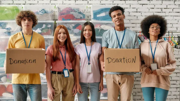 Молодая группа добровольцев улыбается в камеру, держит коробки для пожертвований, счастливая команда работает на благотворительность, жертвует одежду нуждающимся — стоковое фото