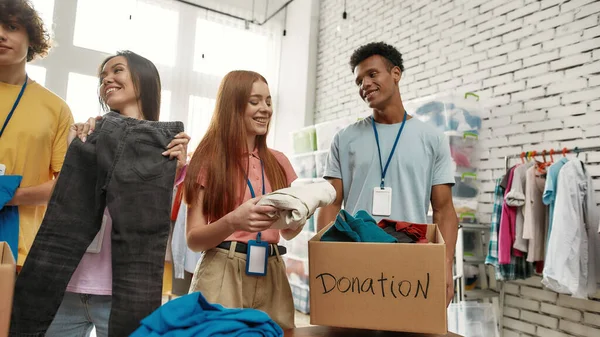 段ボール箱の中に服を詰めながら、幸せの若いボランティアたち、屋内で寄付プロジェクトに取り組む多様なチーム — ストック写真