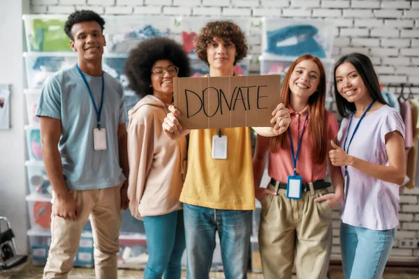 Grupo de diversos jóvenes voluntarios sonriendo a la cámara mientras están de pie en la oficina de la organización benéfica, Guy sosteniendo la tarjeta con Donar letras — Foto de Stock