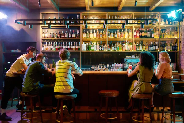 Vista traseira de três tipos a beber cerveja, a olhar para mulheres, duas namoradas sentadas no balcão do bar. Amigos que passam o tempo no clube noturno, restaurante — Fotografia de Stock