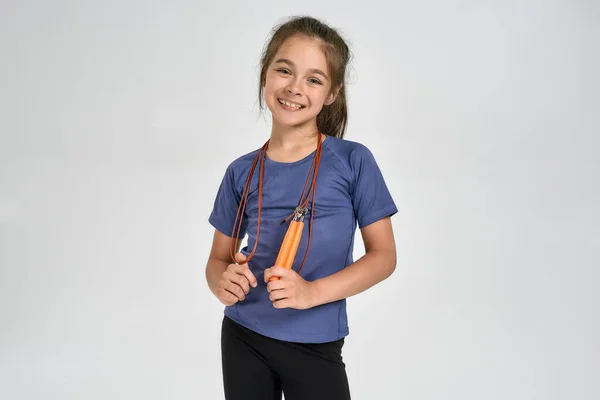 Pequeña niña deportiva en ropa deportiva sonriendo a la cámara, de pie con una cuerda de salto mientras posa aislada sobre un fondo blanco — Foto de Stock