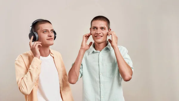 Gozando de lista de reprodução favorita. Dois jovens irmãos gêmeos caucasianos em uso casual ouvindo música em fones de ouvido e fones de ouvido sem fio — Fotografia de Stock