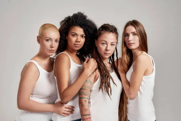 Ritratto di quattro giovani donne diverse che indossano camicie bianche guardando la macchina fotografica mentre posano insieme isolate su sfondo grigio — Foto Stock