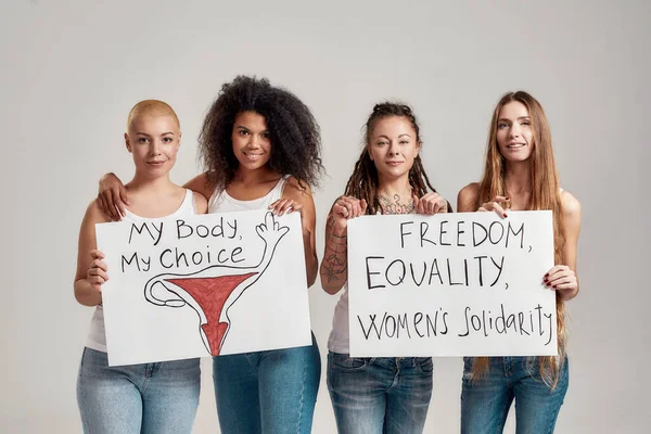 Quattro donne felici e diverse che guardano la macchina fotografica mentre tengono, in piedi con striscioni tra le mani isolati su sfondo grigio. Concetto di femminismo e uguaglianza — Foto Stock