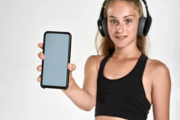 Portrait of cute sportive girl child in headphones, looking at camera, showing smartphone with blank screen, standing isolated over white background — Stock Photo, Image
