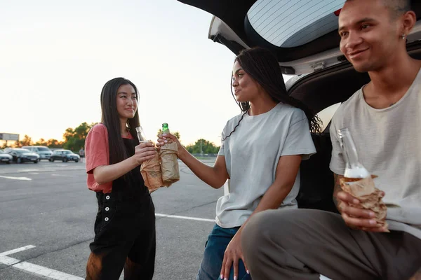 Un grupo de tres amigos de piel oscura de diferente género se divierten juntos afuera en un sitio de estacionamiento bebiendo cerveza sonriéndose entre sí con un maletero abierto — Foto de Stock