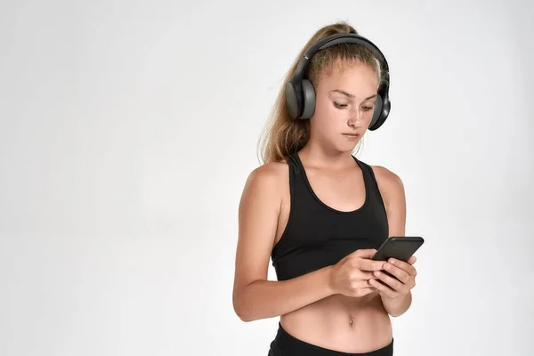 Retrato de una linda niña deportiva que usa auriculares, usando un teléfono inteligente mientras posa aislado sobre un fondo blanco — Foto de Stock