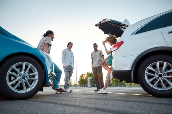 A group of four young well-dressed people speaking to each other standing together outside on a parking site with their cars around