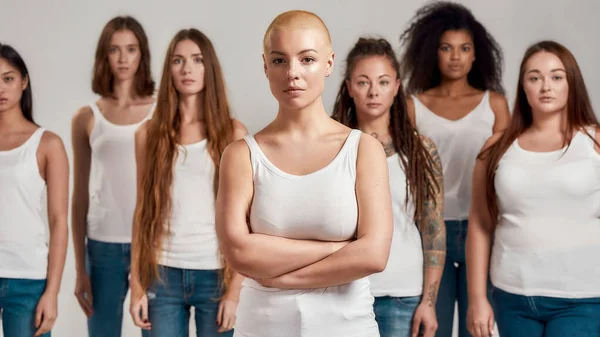 Retrato de bela jovem caucasiana com a cabeça raspada em camisa branca posando com os braços cruzados. Grupo de mulheres diversas de pé isolado sobre fundo cinza — Fotografia de Stock