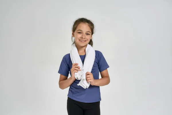 Spor kıyafetli küçük bir kız çocuğu kameraya gülümsüyor, boynuna havlu sarıp beyaz arka planda tek başına duruyor. — Stok fotoğraf