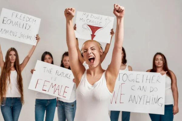Ung kaukasisk kvinna med rakat huvud i vit skjorta höjde armarna och såg entusiastisk ut. Grupp av olika kvinnor som håller protestbanners för kvinnlig makt och rättigheter i bakgrunden — Stockfoto