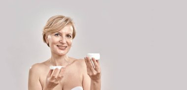 Kameraya gülümseyen orta yaşlı güzel bir kadının portresi. Elinde nemlendirici bir yüz kremi tutarken gri arka planda izole bir şekilde poz veriyor.