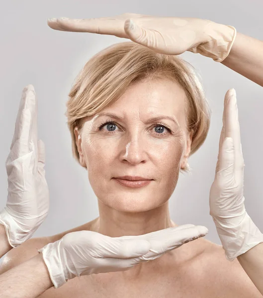 Nahaufnahme Porträt einer attraktiven Frau mittleren Alters, die in die Kamera blickt. Kosmetikerin schafft einen Rahmen um das weibliche Gesicht mit Händen in Handschuhen vor grauem Hintergrund — Stockfoto