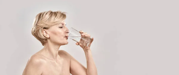 在灰蒙蒙的背景下，一个漂亮的中年妇女在杯子里喝着新鲜的冷水时的近照 — 图库照片