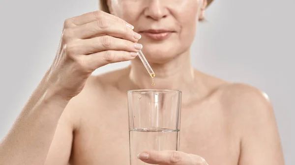 Nahaufnahme der Hand einer Frau mittleren Alters mit Tropfer mit Medikamenten und Glas isoliert über grauem Hintergrund — Stockfoto