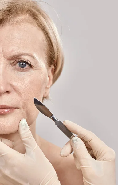 Ujęcie twarzy kobiety w średnim wieku i skalpel medyczny w rękach lekarzy odizolowanych na szarym tle — Zdjęcie stockowe