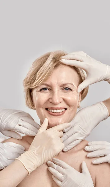 Beauty portrait of middle aged woman smiling at camera. Beautician hands in gloves checking female face skin isolated against grey background — Stock Photo, Image