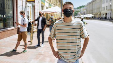 Kameraya bakan maske takan Asyalı bir gencin portresi. Arkaplanda Coronavirus tecridi sırasında insanlar siparişlerini toplanma noktasından alıyorlar.