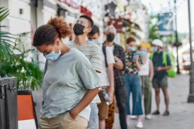 Maske takan genç Afro-Amerikalı kadın bekliyor, diğer insanlarla aynı sırada duruyor, tecrit sırasında toplanma noktasından sipariş almak için sosyal mesafeye saygı duyuyor.